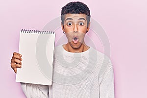 Young african amercian man holding notebook scared and amazed with open mouth for surprise, disbelief face