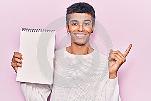 Young african amercian man holding notebook smiling happy pointing with hand and finger to the side