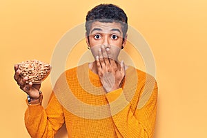 Young african amercian man holding peanuts covering mouth with hand, shocked and afraid for mistake