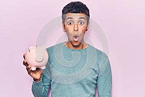 Young african amercian man holding piggy bank scared and amazed with open mouth for surprise, disbelief face