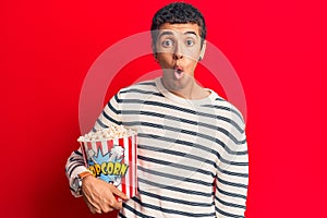 Young african amercian man holding popcorn scared and amazed with open mouth for surprise, disbelief face