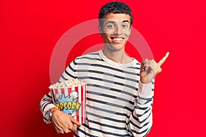 Young african amercian man holding popcorn smiling happy pointing with hand and finger to the side