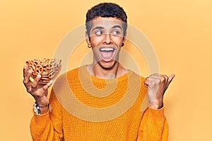 Young african amercian man holding pretzels pointing thumb up to the side smiling happy with open mouth