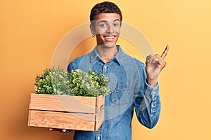 Young african amercian man holding wooden pot with plant smiling happy pointing with hand and finger to the side