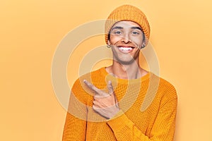 Young african amercian man wearing wool cap smiling cheerful pointing with hand and finger up to the side