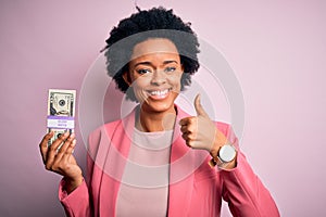 Young African American afro businesswoman with curly hair holding dollars banknotes happy with big smile doing ok sign, thumb up