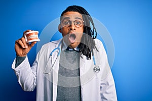 Young african american afro dentist doctor man wearing stethoscope holding teeth scared in shock with a surprise face, afraid and