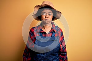 Young African American afro farmer woman with curly hair wearing apron and hat puffing cheeks with funny face