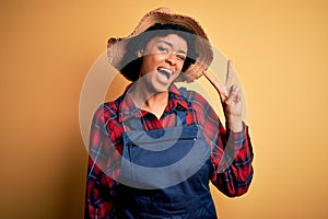 Young African American afro farmer woman with curly hair wearing apron and hat smiling with happy face winking at the camera doing