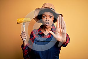 Young African American afro farmer woman with curly hair wearing apron holding cob corn with open hand doing stop sign with