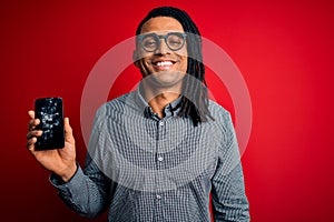 Young african american afro man holding broken smartphone showing craked screen with a happy face standing and smiling with a