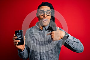 Young african american afro man holding broken smartphone showing craked screen with surprise face pointing finger to himself