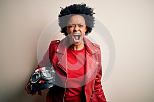 Young African American afro motorcyclist woman with curly hair holding motorcycle helmet angry and mad screaming frustrated and