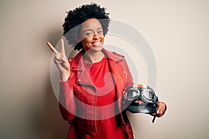 Young African American afro motorcyclist woman with curly hair holding motorcycle helmet smiling with happy face winking at the