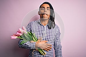 Young african american afro romantic man with dreadlocks holding bouquet of pink tulips making fish face with lips, crazy and