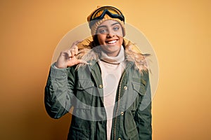 Young african american afro skier girl wearing snow sportswear and ski goggles smiling and confident gesturing with hand doing