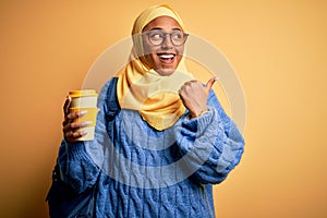 Young African American afro student woman wearing muslim hijab and glasses drinking coffee pointing and showing with thumb up to