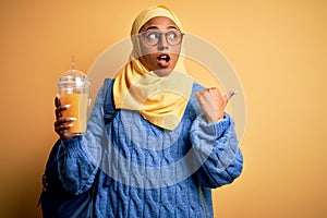 Young African American afro tourist woman wearing muslim hijab drinking orange juice pointing and showing with thumb up to the