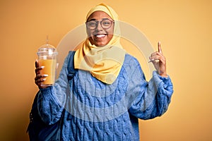 Young African American afro tourist woman wearing muslim hijab drinking orange juice surprised with an idea or question pointing