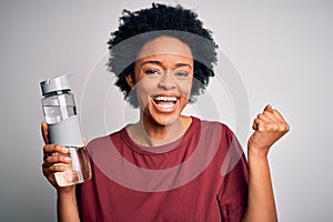 Young African American afro woman with curly hair drinking bottle of water for refreshment screaming proud and celebrating victory