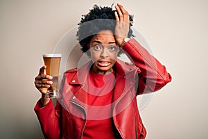Young African American afro woman with curly hair drinking glass of beer with alcohol stressed with hand on head, shocked with