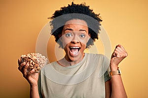 Young African American afro woman with curly hair holding bowl with salty peanuts screaming proud and celebrating victory and
