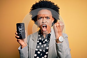 Young African American afro woman with curly hair holding cracked broken smartphone annoyed and frustrated shouting with anger,