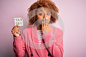 Young African American afro woman with curly hair holding papaer with tax day message cover mouth with hand shocked with shame for