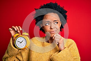 Young African American afro woman with curly hair holding vintage alarm clock serious face thinking about question, very confused