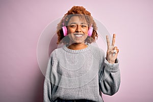 Young African American afro woman with curly hair listening to music using pink headphones smiling with happy face winking at the