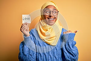 Young African American afro woman wearing muslim hijab holding love reminder paper message pointing and showing with thumb up to