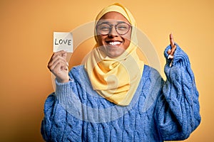 Young African American afro woman wearing muslim hijab holding love reminder paper message surprised with an idea or question