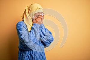 Young African American afro woman wearing muslim hijab over isolated yellow background with sad expression covering face with