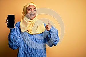 Young African American afro woman wearing muslim hijab showing smartphone happy with big smile doing ok sign, thumb up with