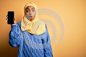 Young African American afro woman wearing muslim hijab showing smartphone scared in shock with a surprise face, afraid and excited