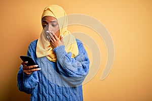 Young African American afro woman wearing muslim hijab using smartphone cover mouth with hand shocked with shame for mistake,