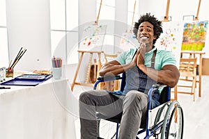 Young african american artist man sitting on wheelchair at art studio praying with hands together asking for forgiveness smiling