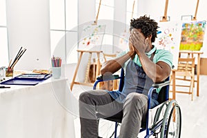 Young african american artist man sitting on wheelchair at art studio with sad expression covering face with hands while crying