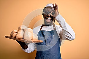 Young african american baker man wearing apron holding tray with homemade bread with happy face smiling doing ok sign with hand on