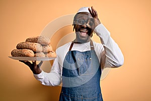 Young african american bakery man holding tray with healthy wholemeal bread with happy face smiling doing ok sign with hand on eye
