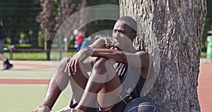 Young african american basketball player eating protein bar, resting after training at streetball court, leaning tree