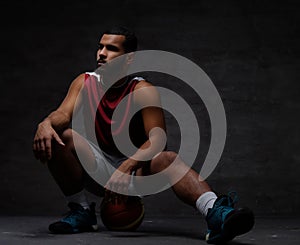 Young African-American basketball player in sportswear sitting on a ball on a dark background.