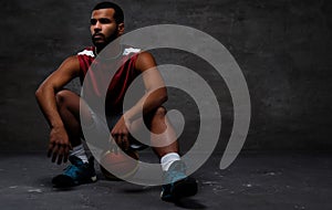 Young African-American basketball player in sportswear sitting on a ball on a dark background.