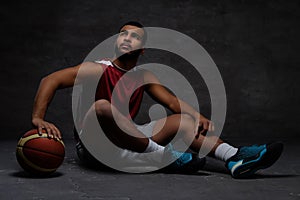 Young African-American basketball player in sportswear sitting on a floor with a ball on a dark background.