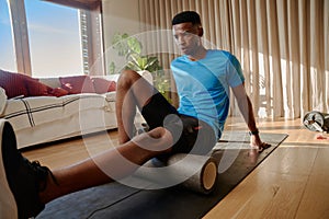 Young African American Black male stretching his hamstring using a foam roller on an exercise mat, working out at home