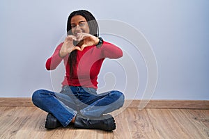Young african american with braids sitting on the floor at home smiling in love doing heart symbol shape with hands