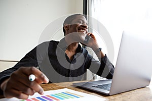 Young african american business man sitting at desk with laptop talking on cellphone