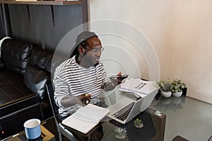 Young African American business man working with laptop and document on desk and talking on mobile at home