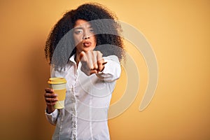 Young african american business woman with afro hair drinking coffee from take away cup pointing with finger to the camera and to