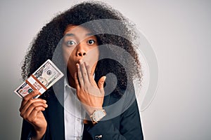 Young african american business woman with afro hair holding a bunch of cash dollars banknotes cover mouth with hand shocked with